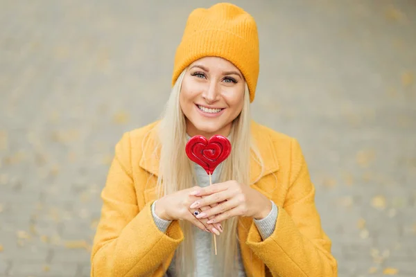 Saint Valentine\'s day concept. Fashion portrait blond young woman in yellow coat having fun with red lollipop heart over street background.