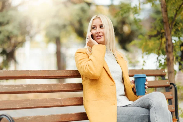 Young blond woman sitting in the city park and  drinking coffee to go, using mobile phone