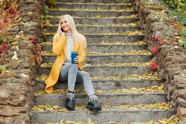 Young blond woman sitting in the city park and  drinking coffee to go, using mobile phone