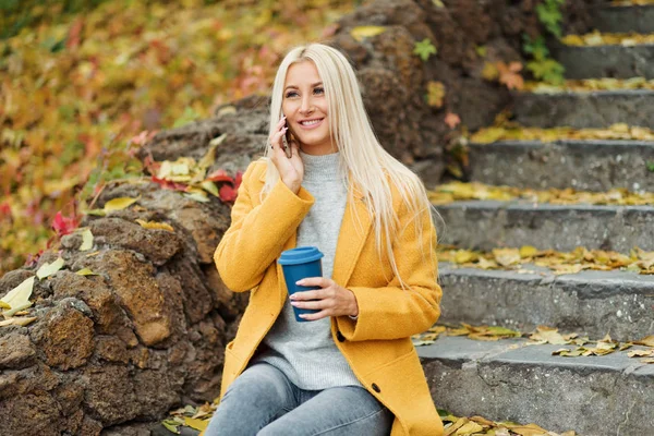 Young blond woman sitting in the city park and  drinking coffee to go, using mobile phone