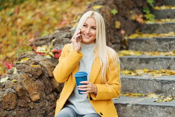 Young blond woman sitting in the city park and  drinking coffee to go, using mobile phone