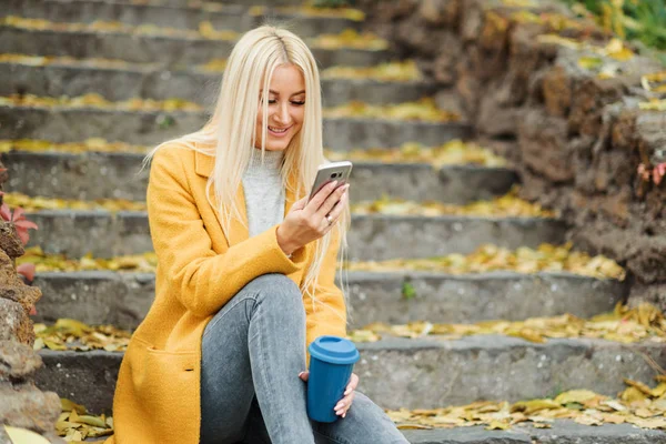 Young blond woman sitting in the city park and  drinking coffee to go, using mobile phone