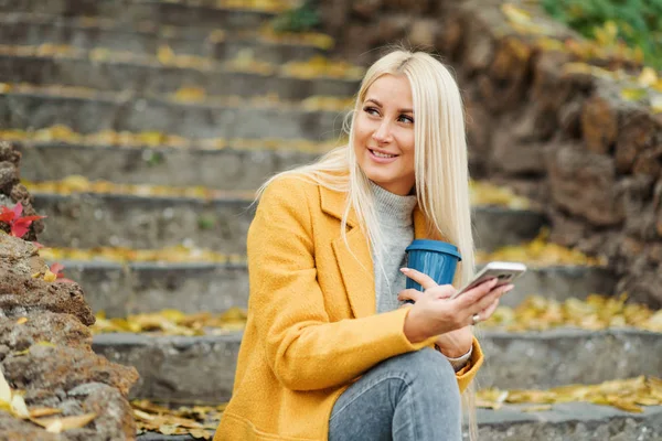 Young blond woman sitting in the city park and  drinking coffee to go, using mobile phone