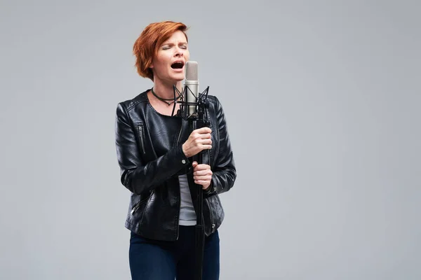 Portrait of rock singer wearing leather jacket and keeping static mic, sings a song loudly on grey background. Concept of rock music and rave