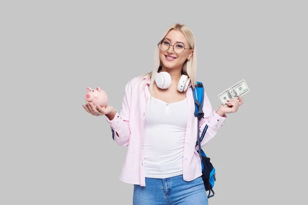 Cost of college education. Portrait of a happy  female student  with piggy bank and cash money in  hands. The girl saves money for training in high school .Neutral grey background. Space for text