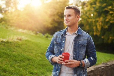 Parkta paket servisi olan restoran kahve ile ayakta, plesantly kameraya gülümseyen genç Hipster adam. Mavi Kot ceket mutlu kaygısız yakışıklı adam