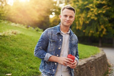 Parkta paket servisi olan restoran kahve ile ayakta, plesantly kameraya gülümseyen genç Hipster adam. Mavi Kot ceket mutlu kaygısız yakışıklı adam