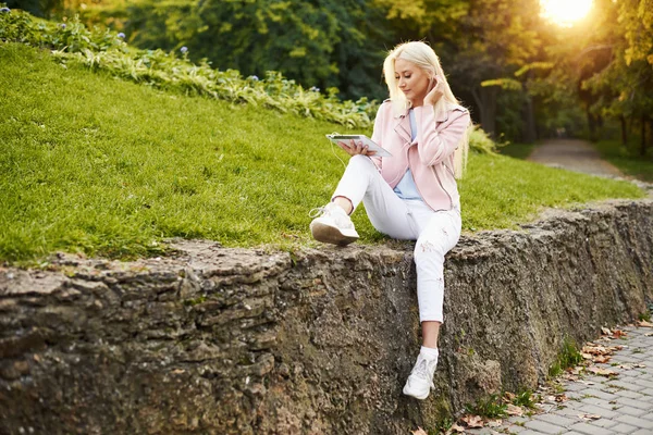 Smiling handsome woman listening relaxing music In the headphones  with her mobile phone or tablet at the park. A student listens to an online lecture and prepares for exams. girl enjoying loneliness