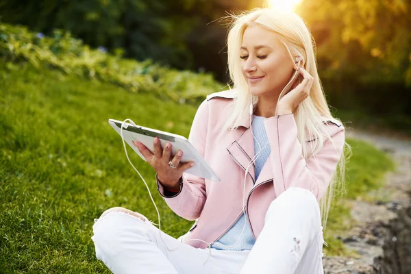 Smiling handsome woman listening relaxing music In the headphones  with her mobile phone or tablet at the park. A student listens to an online lecture and prepares for exams. girl enjoying loneliness