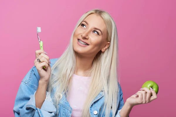 Diş hekimi ve ortodontist kavramı. Temizlik ve diş fırçalama diş fırçası kullanarak diş telleri ile genç sarışın gülümseyen kadın. Bir yeşil elma ısırmaya çalışıyor. Pembe bir arka plan