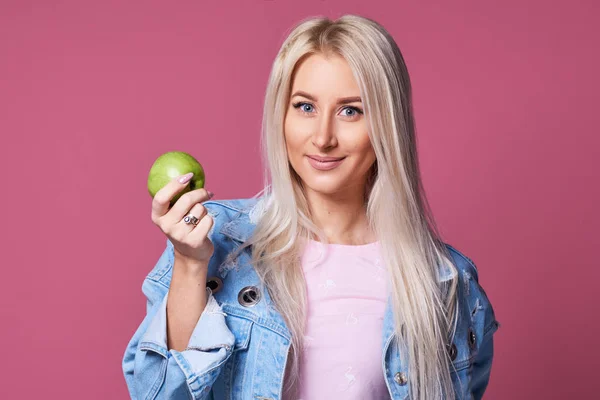 A young girl blonde holds in her hand a green apple on a pink background. Solid food and braces