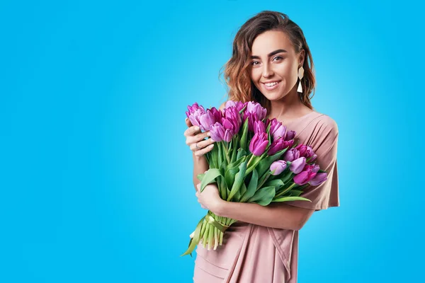 Üzerinde mavi arka plan izole mor Lale buketi tutan genç mutlu gülümseyen Kızıl saçlı kadın. Kadın 8 Mart gününde veya doğum günü onuruna şenlikli buket