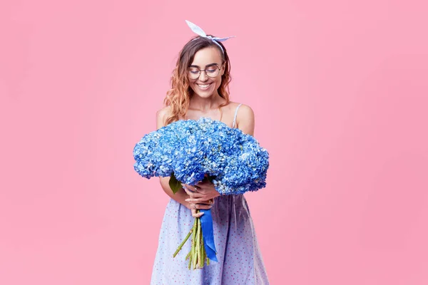 Cheerful young lady in retro round glasses being excited to get bouquet of blue spring flowers on women's day isolated over pink background. It is looking to the right, space for text