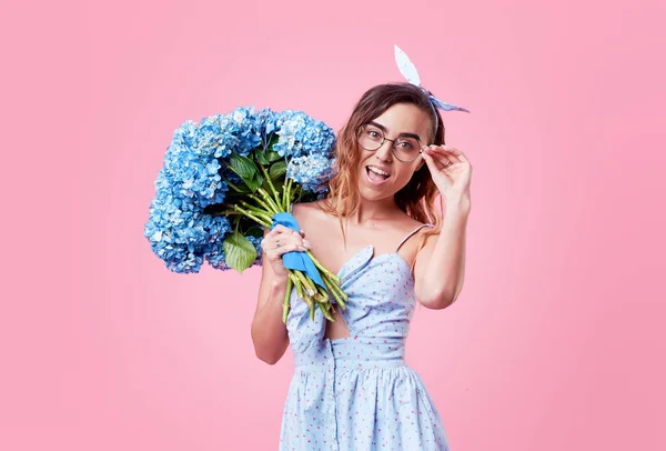 Cheerful young lady in retro round glasses being excited to get bouquet of blue spring flowers on women\'s day isolated over pink background. It is looking to the right, space for text