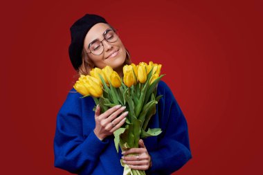 Cheerful young lady in blue clothes, black beret and round glasses being excited to get bouquet of yellow tulips on women's day or birthday isolated over red background. French style girl  clipart