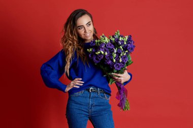 Cheerful young lady in blue clothes, holding and smelling bouquet of colorful tulips on women's day or birthday isolated over red background.  clipart