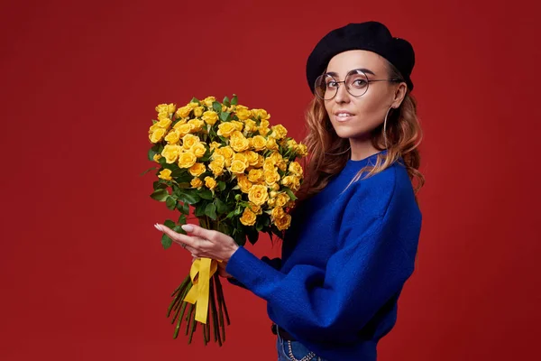 青い服 黒ベレー帽と丸眼鏡女性の日や 赤い背景に分離の誕生日に黄色のバラの花束を得るに興奮している陽気な若い女性 フランス スタイルの女の子 — ストック写真