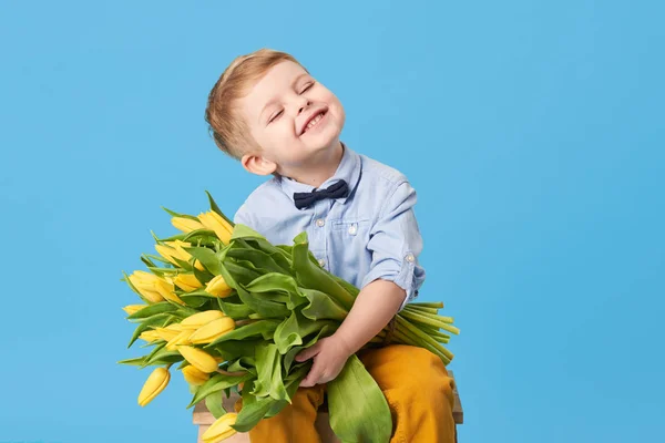 Adorable smiling child with spring flower bouquet looking at camera isolated on blue. Little toddler boy holding yellow tulips as gift for mom