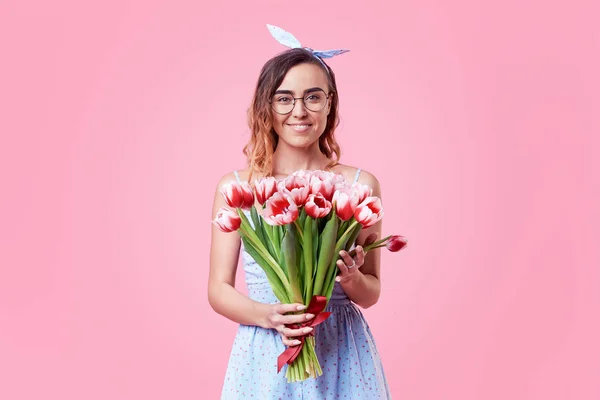 Cheerful young lady in retro round glasses being excited to get bouquet of red spring flowers on women\'s day isolated over pink background. It is looking to the right, space for text