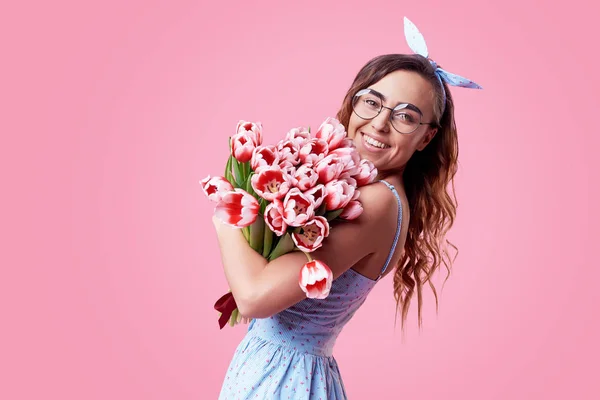 Cheerful young lady in retro round glasses being excited to get bouquet of red spring flowers on women's day isolated over pink background. It is looking to the right, space for text