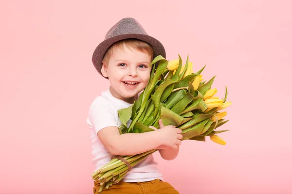 Adorable smiling child with spring flower bouquet looking at camera isolated on pink. Little toddler boy in hat holding yellow tulips as gift for mom. Copy space for text on right side