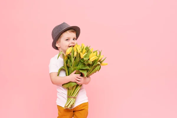 Adorable smiling child with spring flower bouquet looking at camera isolated on pink. Little toddler boy in hat holding yellow tulips as gift for mom