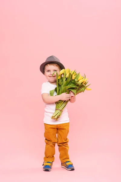 Adorable smiling child with spring flower bouquet looking at camera isolated on pink. Little toddler boy in hat holding yellow tulips as gift for mom