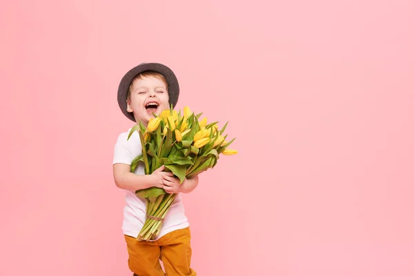 Adorable smiling child with spring flower bouquet looking at camera isolated on pink. Little toddler boy in hat holding yellow tulips as gift for mom