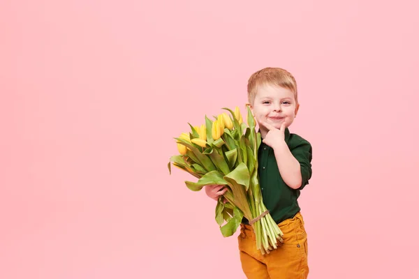 Adorable smiling child with spring flower bouquet looking at camera isolated on pink. Little toddler boy holding yellow tulips as gift for mom. Copy space for text on left side