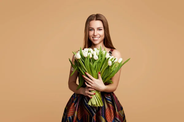 Wow effect. Wonderful flowers on Womens Day. Tender smiling brunet woman poses with white tulips, looks happily, isolated over beige studio background wall. Space for text
