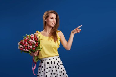 Mükemmel bir hediye. Bayan gün harika çiçekler. İhale gülümseyen brunet kadın renkli Lale ile görünüyor ne mutlu ki, mavi stüdyo arka plan üzerinde poz veriyor. Sağdaki bir parmağını gösterir. boş metin alanı