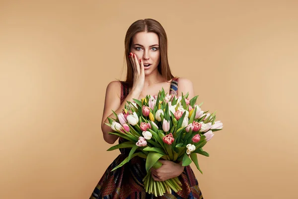 Wow effect. Wonderful spring flowers on Womens Day. Tender surprised brunet woman poses with colorful tulips, looks happily, isolated over beige studio background wall. Space for text