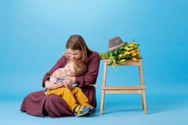 Genç anne ve küçük yürümeye başlayan çocuk oğlu eğleniyor portresi. Neredeyse bir buket bahar sarı çiçek, Lale yatıyor. Aile, sevimli çocuk, sevgi ve mutluluk kavramı