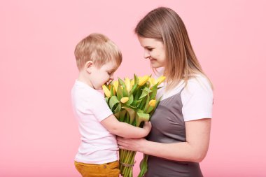 Genç anne küçük oğlu pembe bir arka plan karşı katta oturan sarılmak. Anne bir buket bahar sarı çiçek tutar. Bakım ve ilişkiler ve aile kavramı. Anneler günü