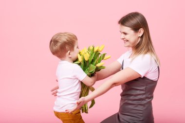 Genç anne küçük oğlu pembe bir arka plan karşı katta oturan sarılmak. Anne bir buket bahar sarı çiçek tutar. Bakım ve ilişkiler ve aile kavramı. Anneler günü