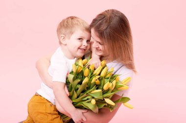 Genç anne küçük oğlu pembe bir arka plan karşı katta oturan sarılmak. Anne bir buket bahar sarı çiçek tutar. Bakım ve ilişkiler ve aile kavramı. Anneler günü
