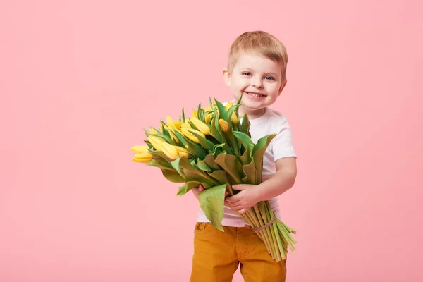 Adorable smiling child with spring flower bouquet looking at camera isolated on pink. Little toddler boy holding yellow tulips as gift for mom. Copy space for text