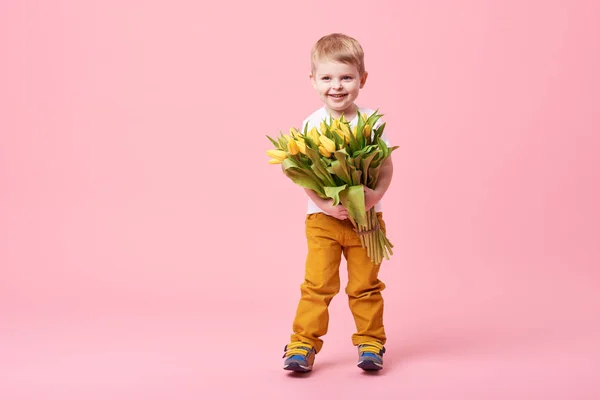 Adorable smiling child with spring flower bouquet looking at camera isolated on pink. Little toddler boy holding yellow tulips as gift for mom. Copy space for text