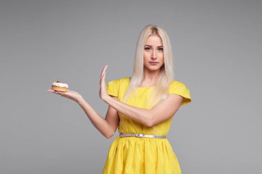 Diyet. Diyet kavramı. Sağlıklı gıda. Sarı elbiseli genç sarışın kadın eller Dur bir lezzetli pasta üzerinde gösterir. Gri arka plan üzerinde izole. Alıcı kafeterya şekerlemeler bölümü