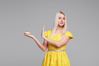 Diyet. Diyet kavramı. Sağlıklı gıda. Sarı elbiseli genç sarışın kadın eller Dur bir lezzetli pasta üzerinde gösterir. Gri arka plan üzerinde izole. Alıcı kafeterya şekerlemeler bölümü