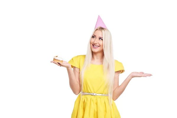 陽気な美しいブロンドの若い女性黄色のドレスと誕生日の帽子やキャップの小さな休暇カップケーキを与えます 白い背景の上の喜びを味わう — ストック写真