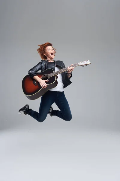 Young woman musician with an acoustic guitar in hand on a gray background. She laughs and jumps high. plays rock and roll loudly. Full-length portrait.