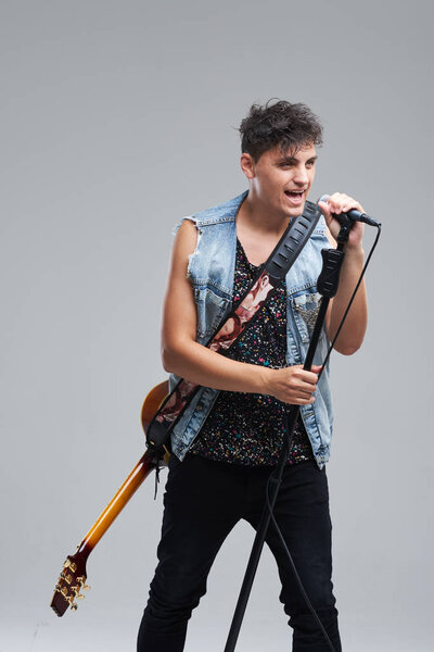 Portrait of rock singer man keeping electric guitar and static acoustic mic, sings a song loudly on grey background. Concept of rock music and rave