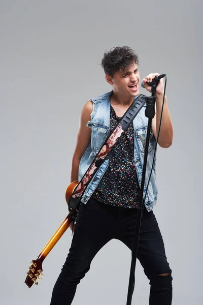 Portrait of rock singer man keeping electric guitar and static acoustic mic, sings a song loudly on grey background. Concept of rock music and rave