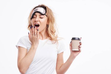 Uykusuz bir gece. Stresli ve yorgun bir uyku maskesi kız bir fincan kahve ve esneme tutar. Beyaz arka planda poz moda dalgalı saçlı beyaz pijama genç bir kadının portresi