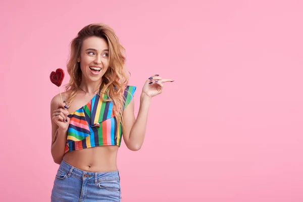 Fashion portrait of pretty sweet young smiling woman having fun with lollipop over pink background. It shows a finger to the right side. Copy space for text