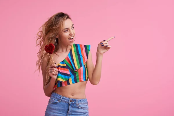 Fashion portrait of pretty sweet young smiling woman having fun with lollipop over pink background. It shows a finger to the right side. Copy space for text