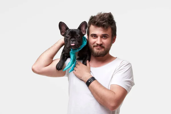 Hipster Sakallı adam tutan ve sevgi ile kollarında güzel bir Fransız Bulldog köpek sarılma ve onunla oynarken, gri bir arka plan karşı
