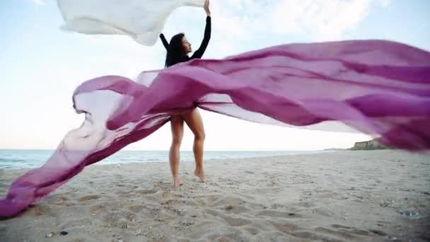 青空スローモーションの下で空の海岸に風に振られた大きなシフォンの作品を保持するスリムな脚のブルネットの女性 — ストック動画