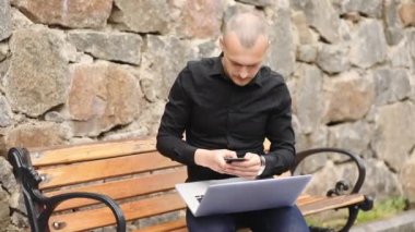 siyah yakışıklı kel hacker kayalık duvarda ahşap bankta oturan akıllı telefon ve dizüstü bilgisayar ile siber saldırı geliştirir